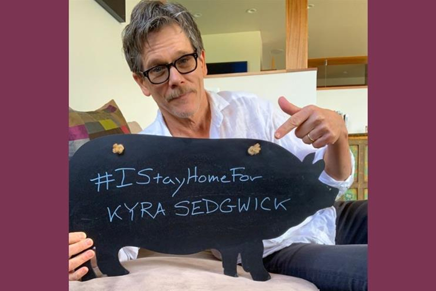 妻(キーラ・セジウィック、女優)の名前とハッシュタグを書いたボードを掲げるケヴィン・ベーコン
